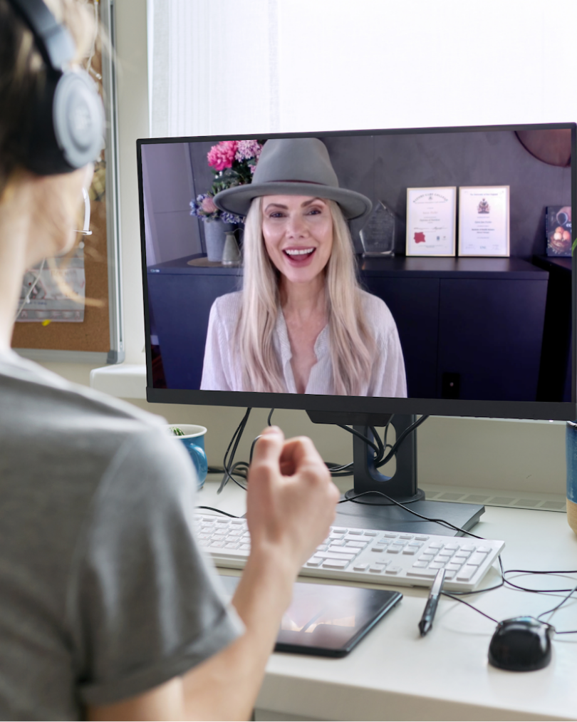 A woman wearing over-ear headphones sits at a desk facing a computer monitor during a video call, with a smiling blonde woman in a gray hat visible on the screen.